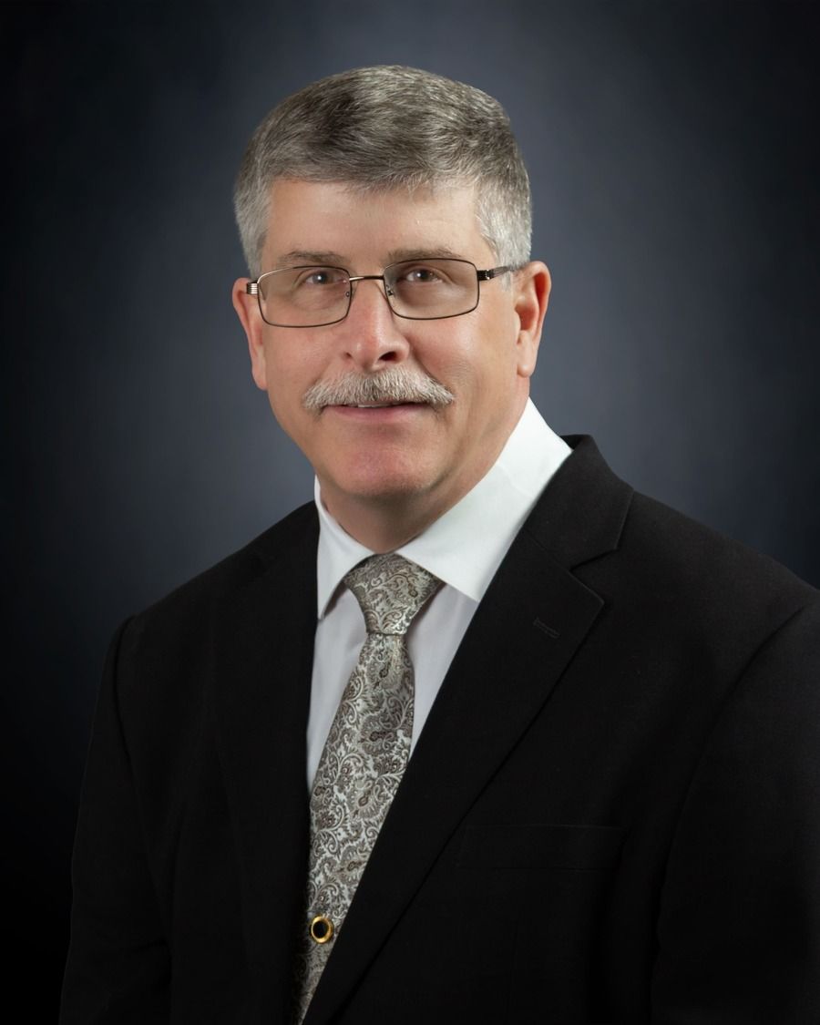 Studio portrait of a man in a dark suit, patterned tie, and glasses against a gray background