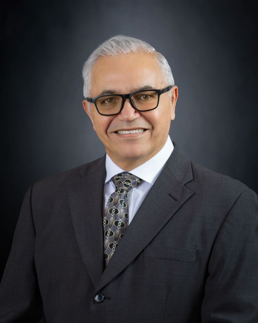 Formal headshot of a smiling person in a dark suit, patterned tie, and glasses against a dark background