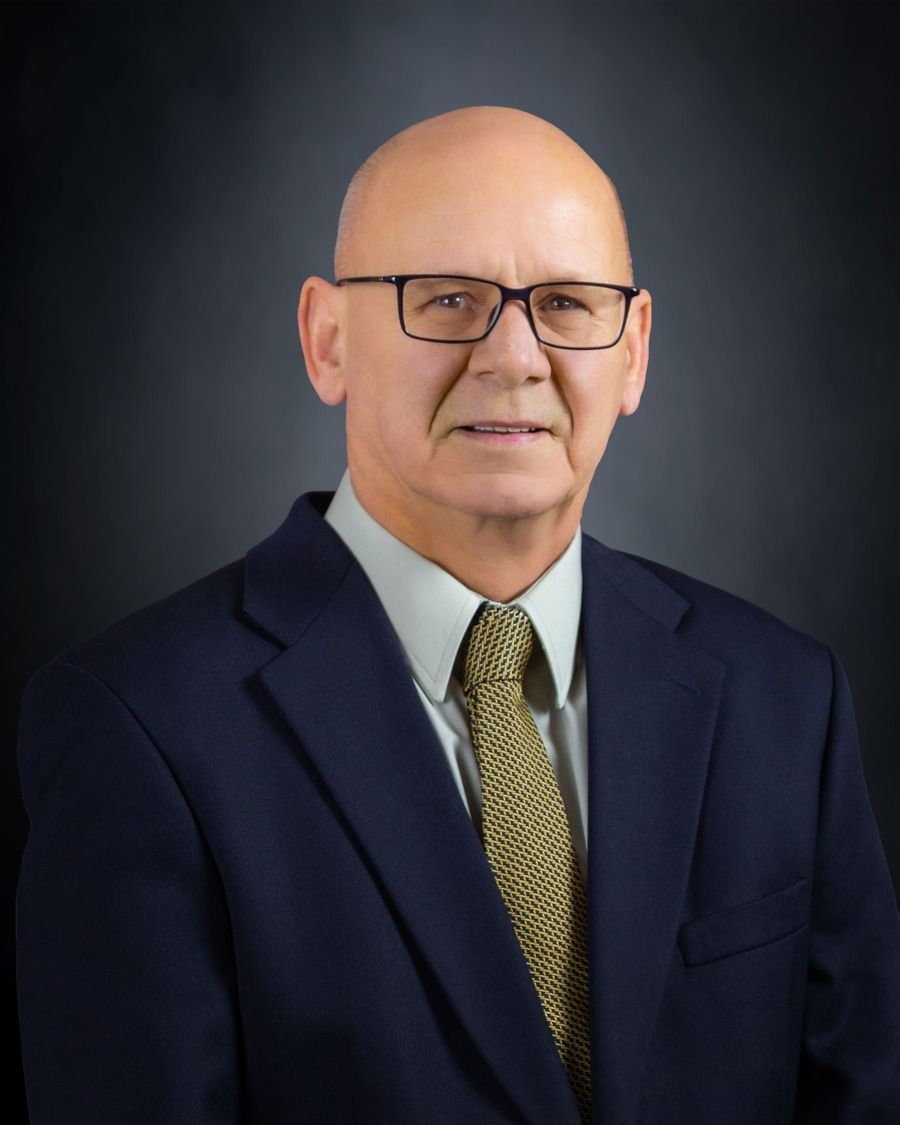 Studio portrait of a man in a dark suit, blue shirt, and patterned tie against a dark background