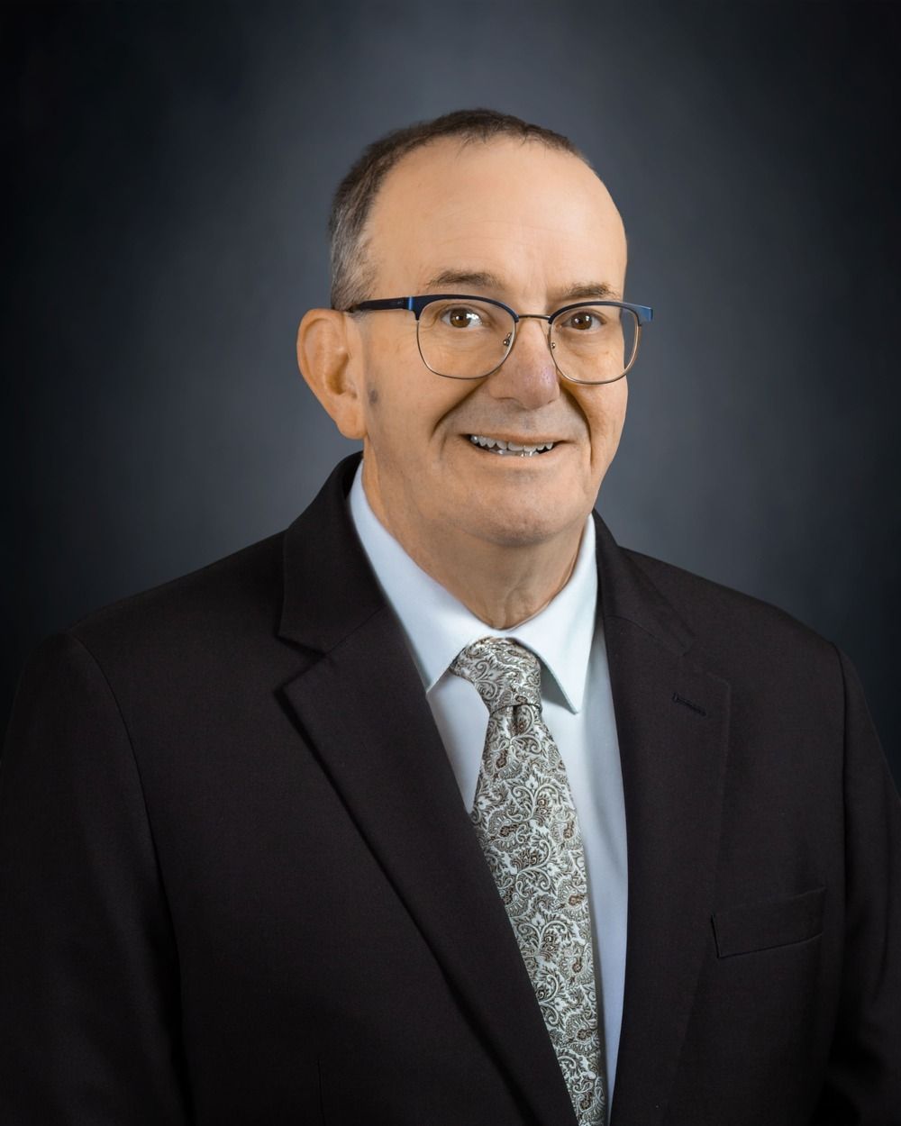 Studio portrait of a man in a dark suit, white shirt, and patterned tie against a gray background