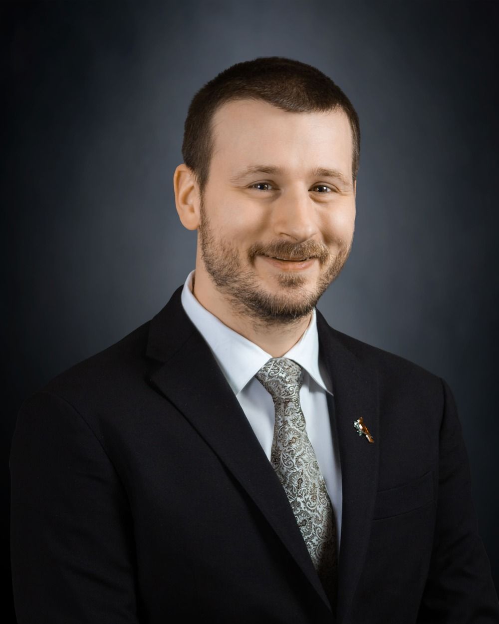 Studio portrait of a man in a dark suit, white shirt, and patterned tie against a dark background