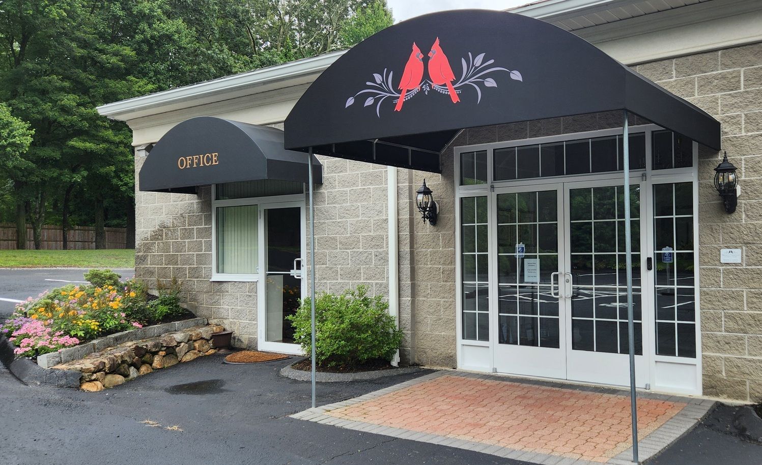 Small stone building entrance with black awning, glass double doors, and flower beds beside a wet driveway