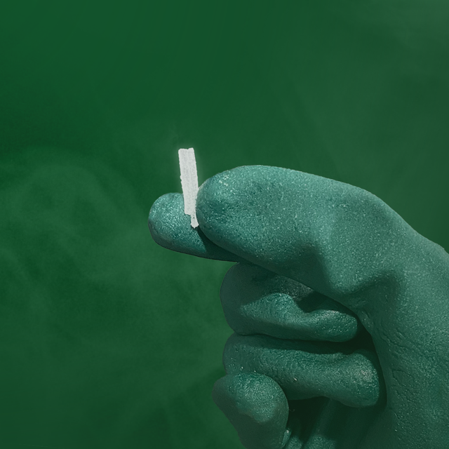 Mão enluvada segurando um pequeno cigarro branco contra um fundo verde.