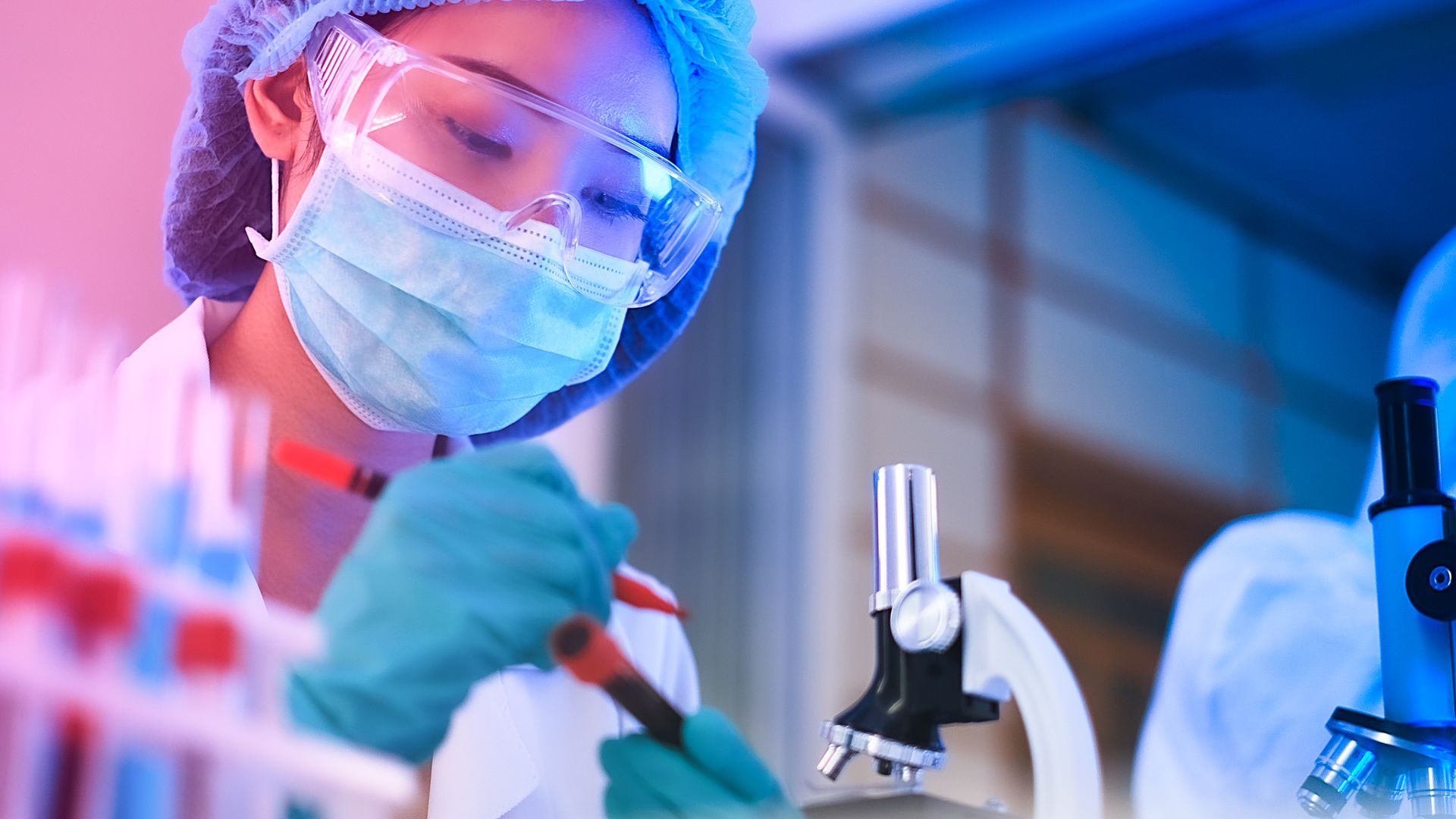 Cientista com óculos de proteção e máscara trabalhando com equipamentos de laboratório sob luz azul.