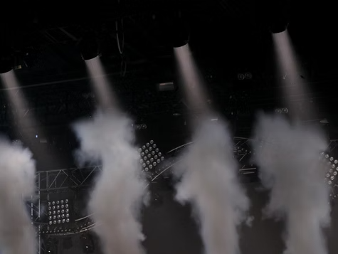 Colunas de fumaça branca se elevam em um palco escuro sob holofotes.