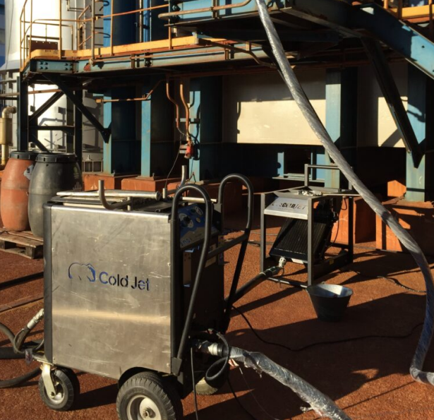 Máquina de corte a frio em uma plataforma industrial enferrujada, com mangueiras e escadas ao fundo.
