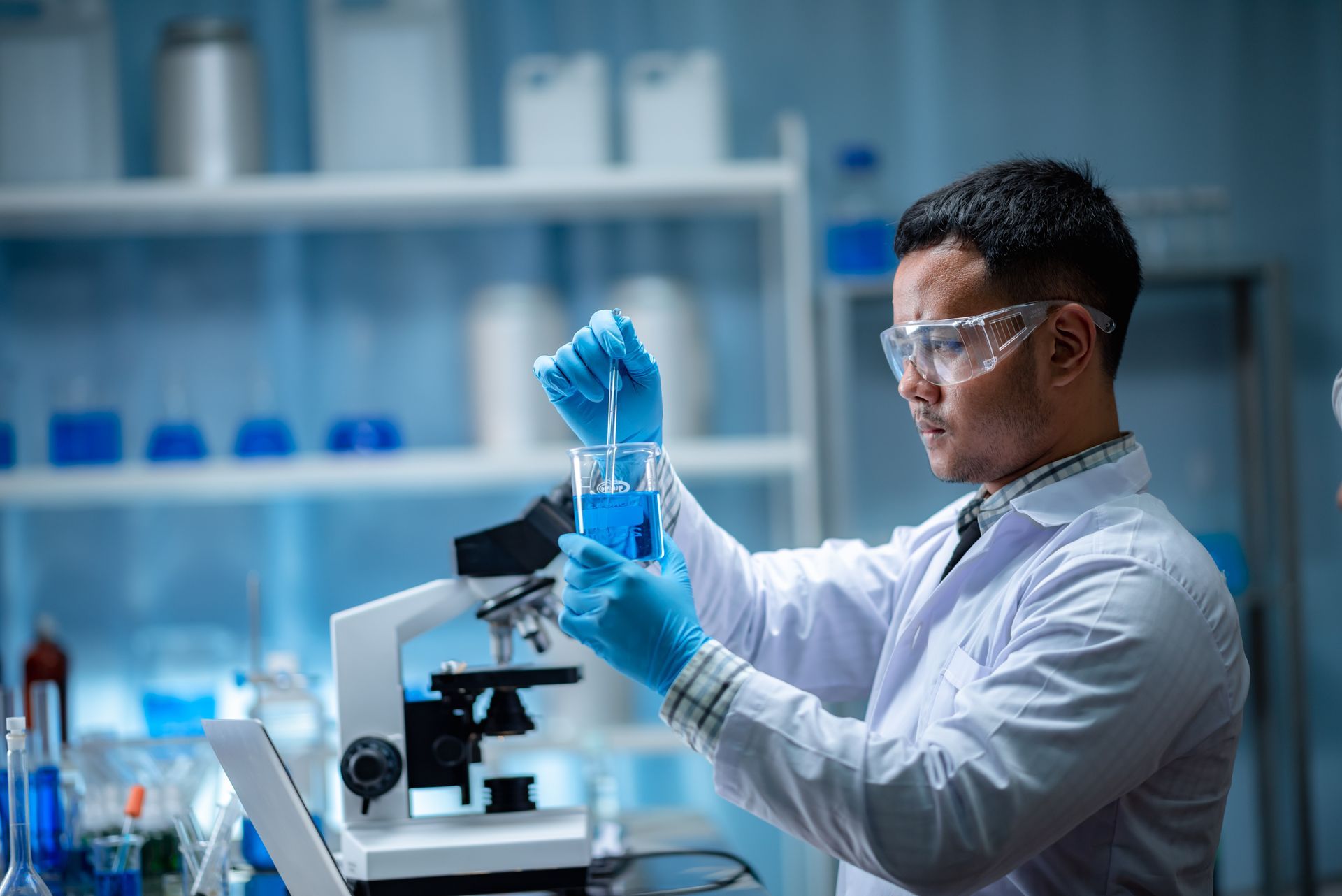 Cientista de jaleco e óculos de proteção manuseando um tubo de ensaio ao lado de um microscópio em um laboratório com iluminação azul.