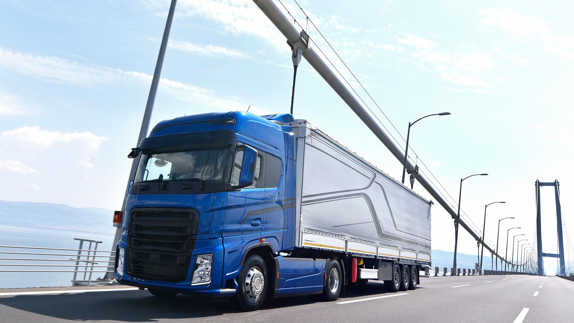 Caminhão semirreboque azul trafegando sobre uma ponte suspensa por cabos sobre a água.