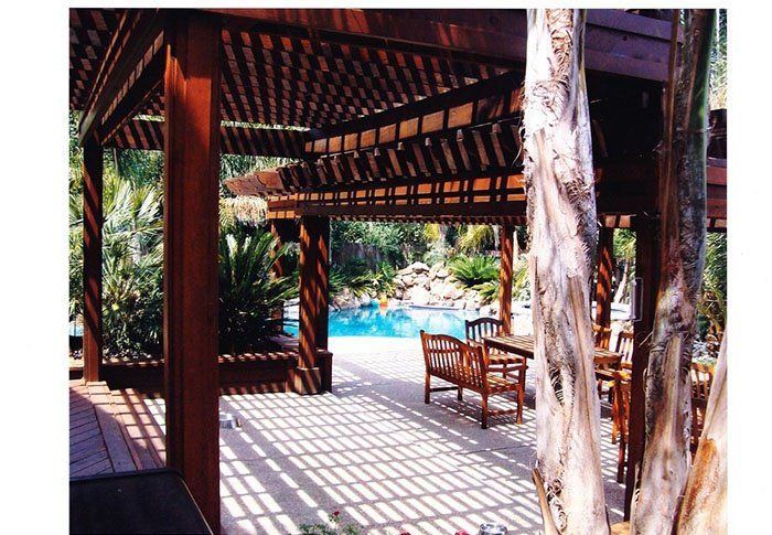 Wooden poolside gazebo with bench, tropical trees, and sunlit blue water beyond
