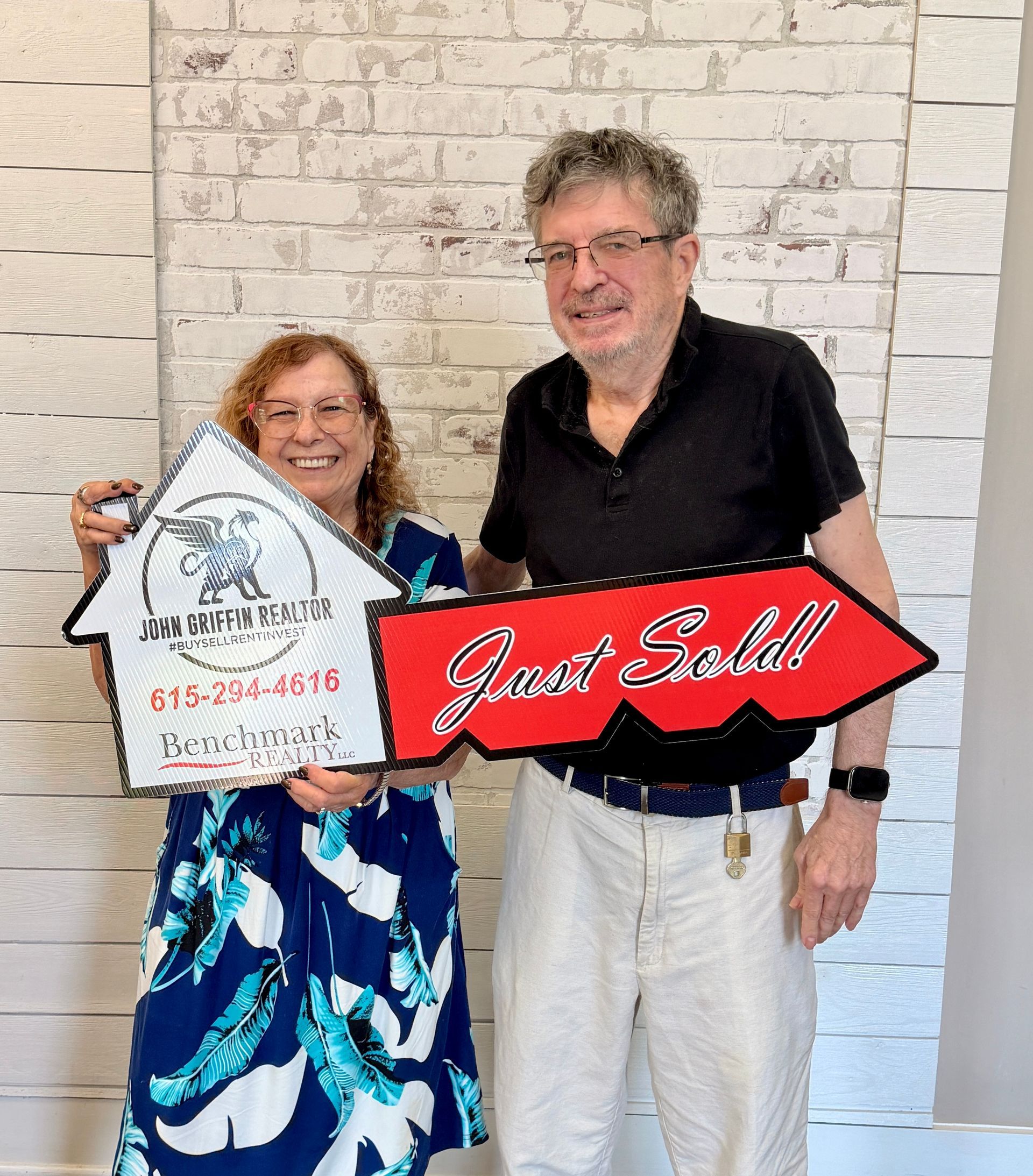 Two people smiling and holding real estate signs in front of a white brick wall.