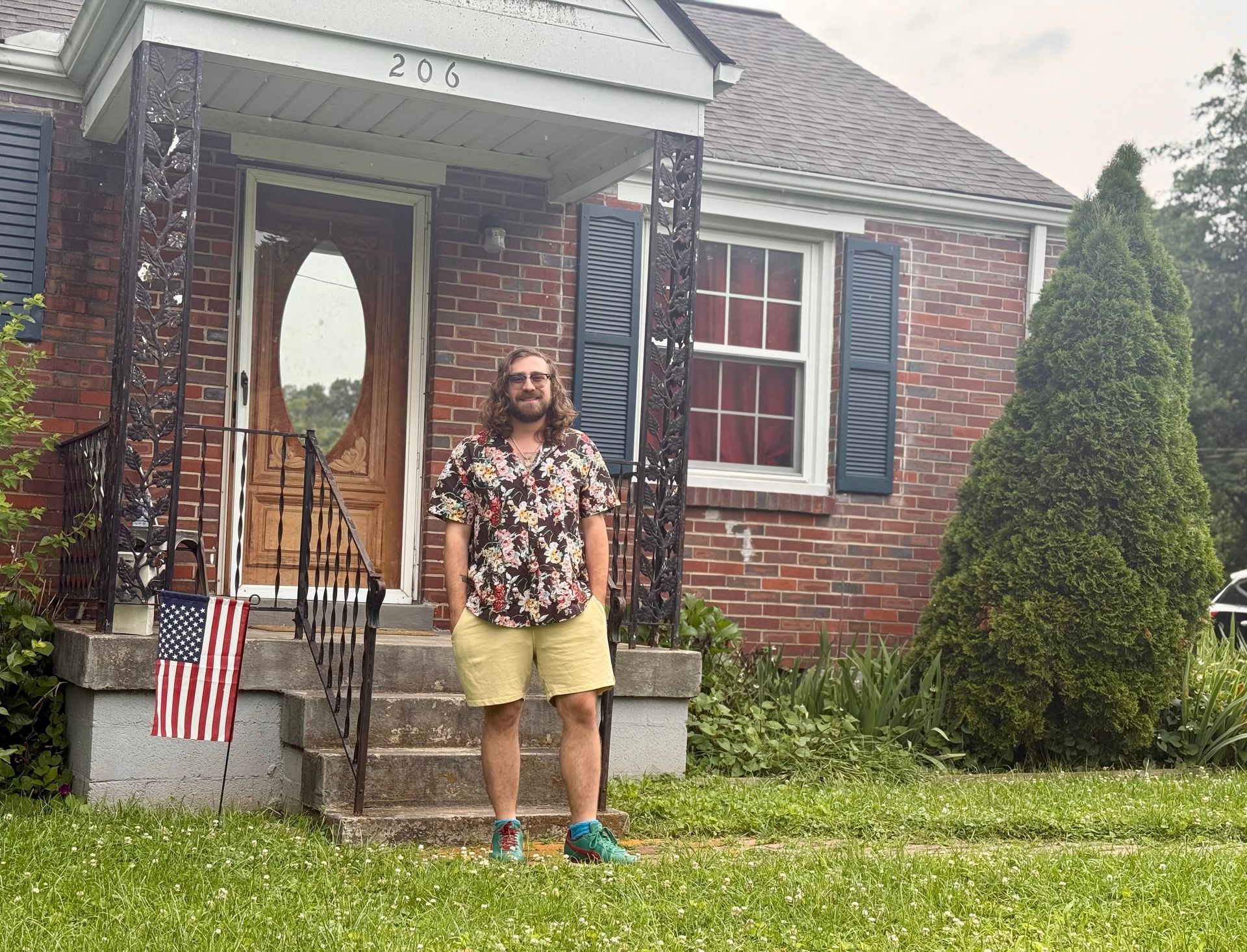 Person standing on a lawn in front of a brick house, wearing a floral shirt, shorts, and green shoes.