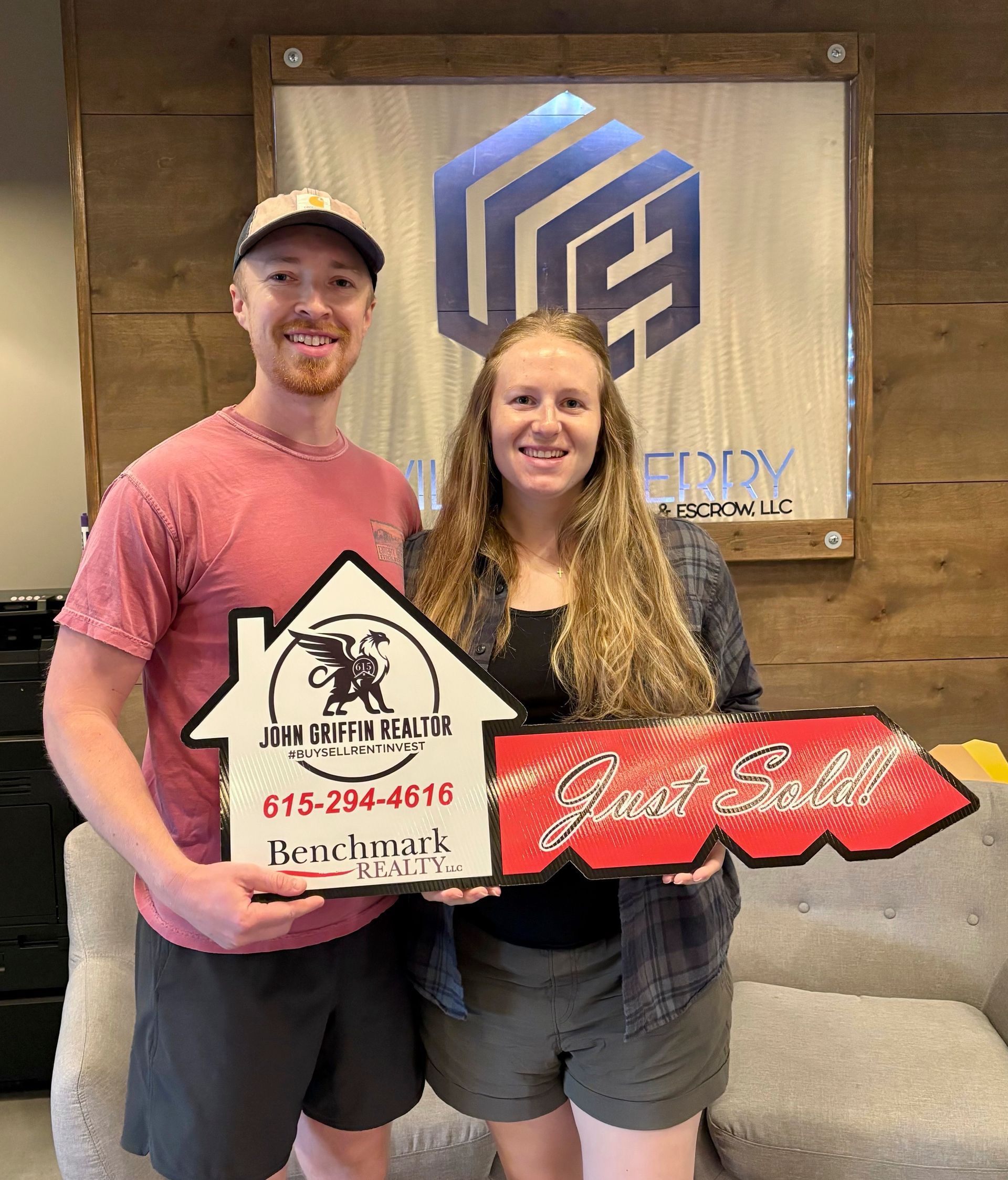Two smiling people holding a house-shaped sign and a red key-shaped sign in front of a logo wall