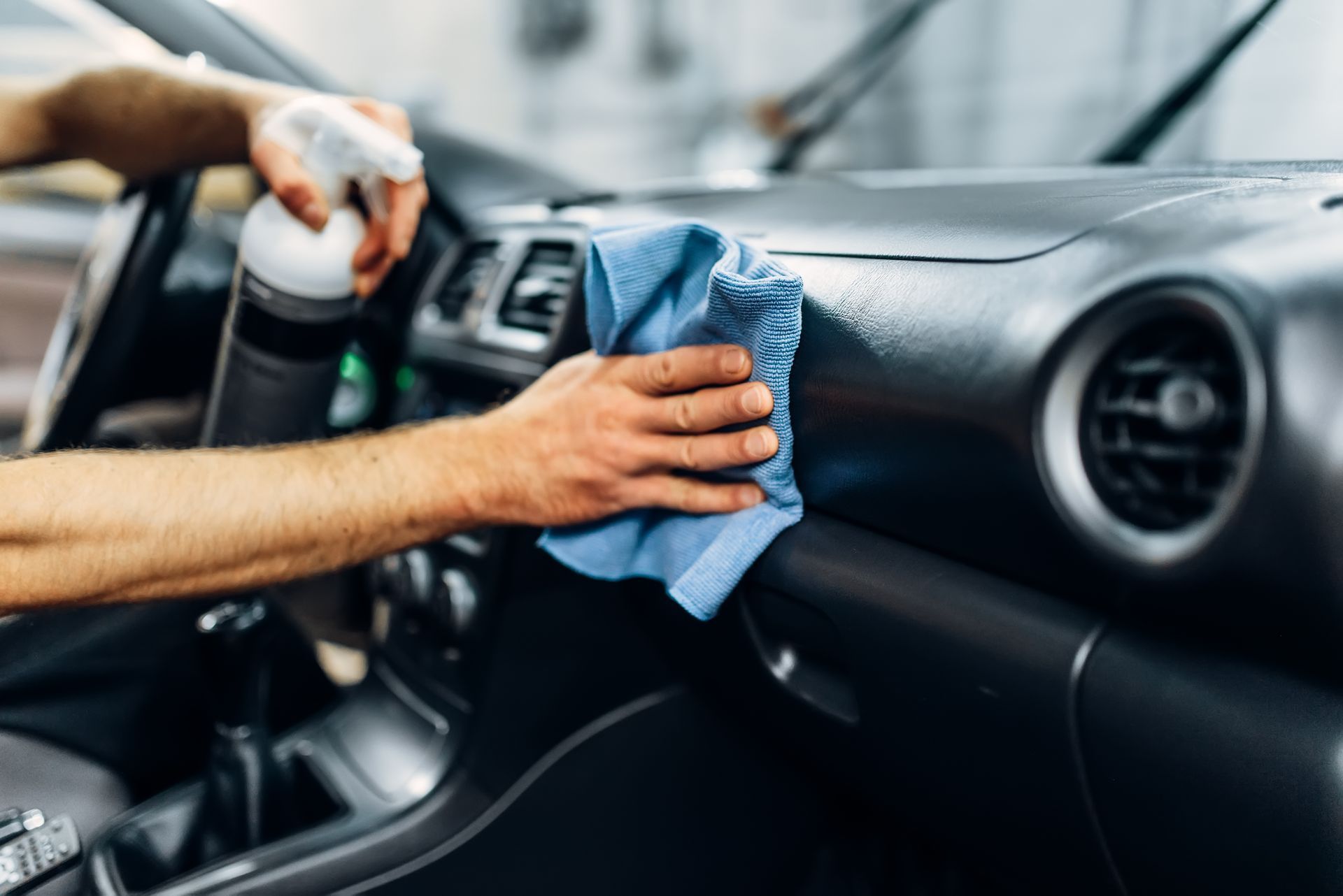 Person cleaning a car dashboard with a blue cloth and spray bottle