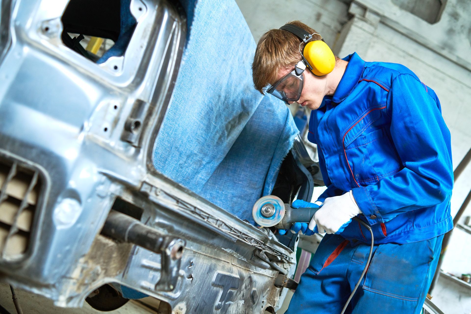 Technician in blue coveralls and yellow earmuffs grinding a metal car body panel in a workshop