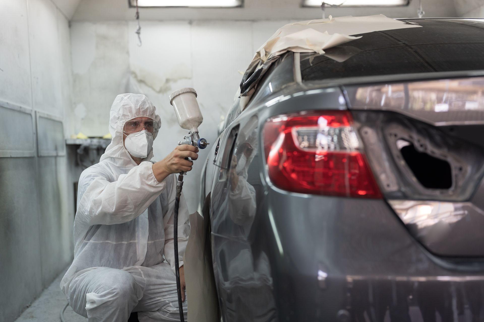 Painter spray-painting a gray car in a body shop, wearing protective coveralls and respirator.