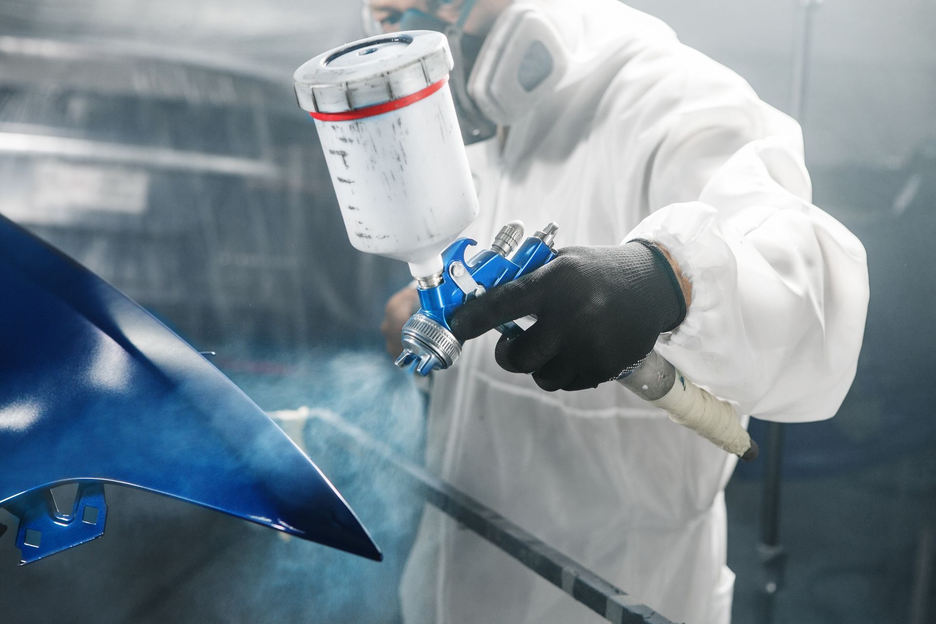 Worker spray-painting a blue car panel in a paint booth with a spray gun