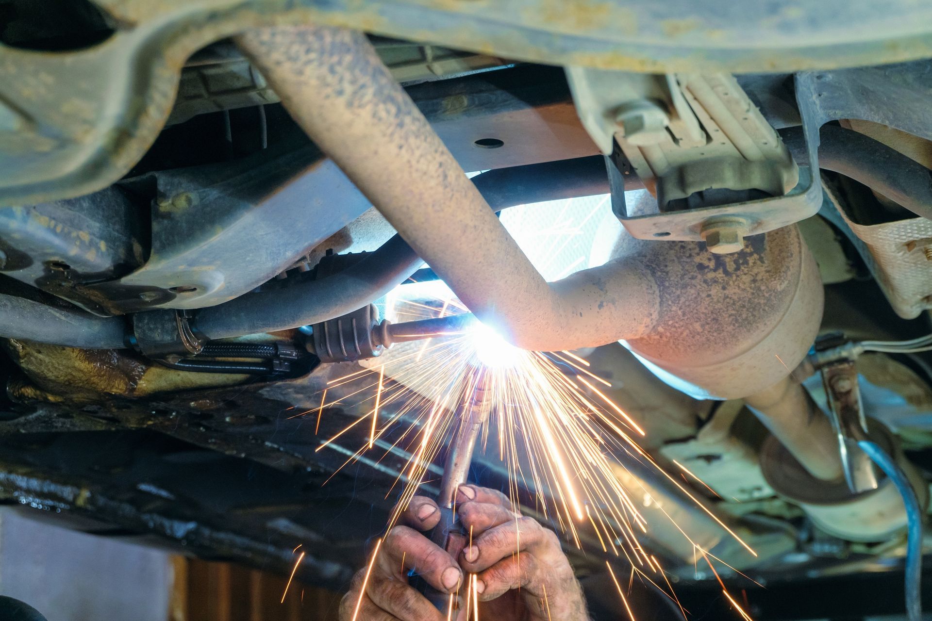 Mechanic welding under a car, with bright sparks near the exhaust system and undercarriage.