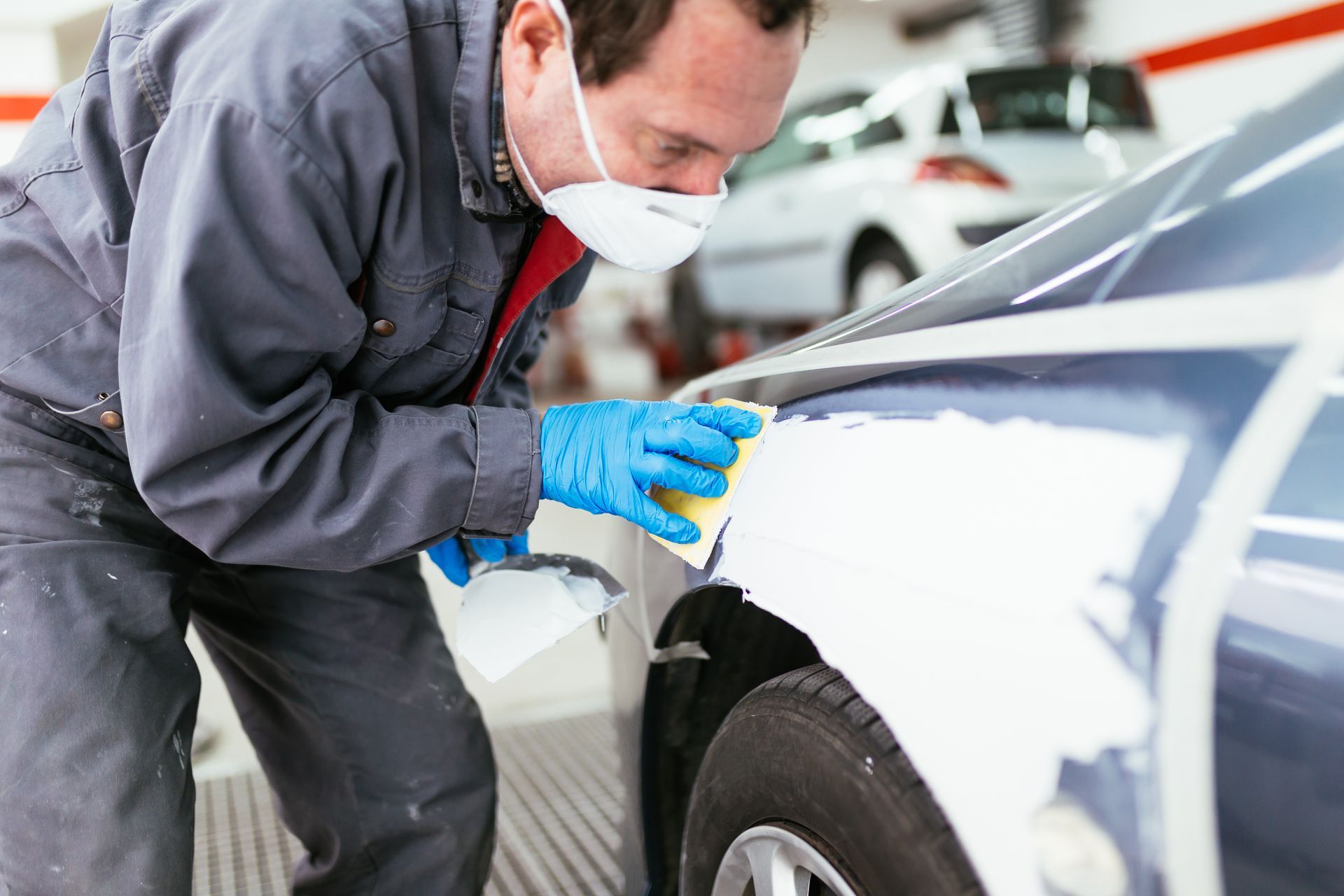 Person wearing a mask and blue gloves painting a white car panel in an auto body shop