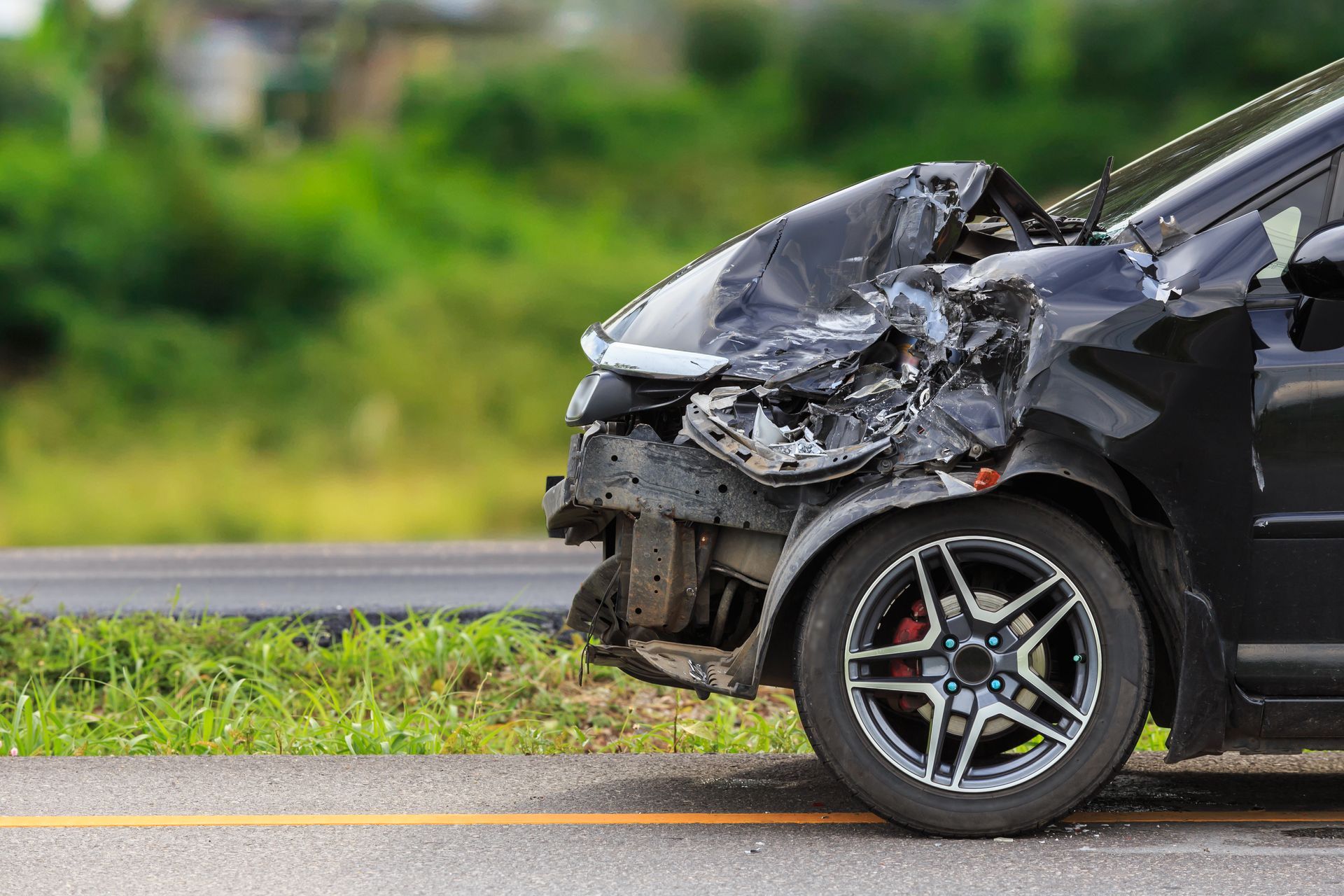 Severely damaged black car front-end after a crash on a roadside, with a bent wheel and smashed hood.