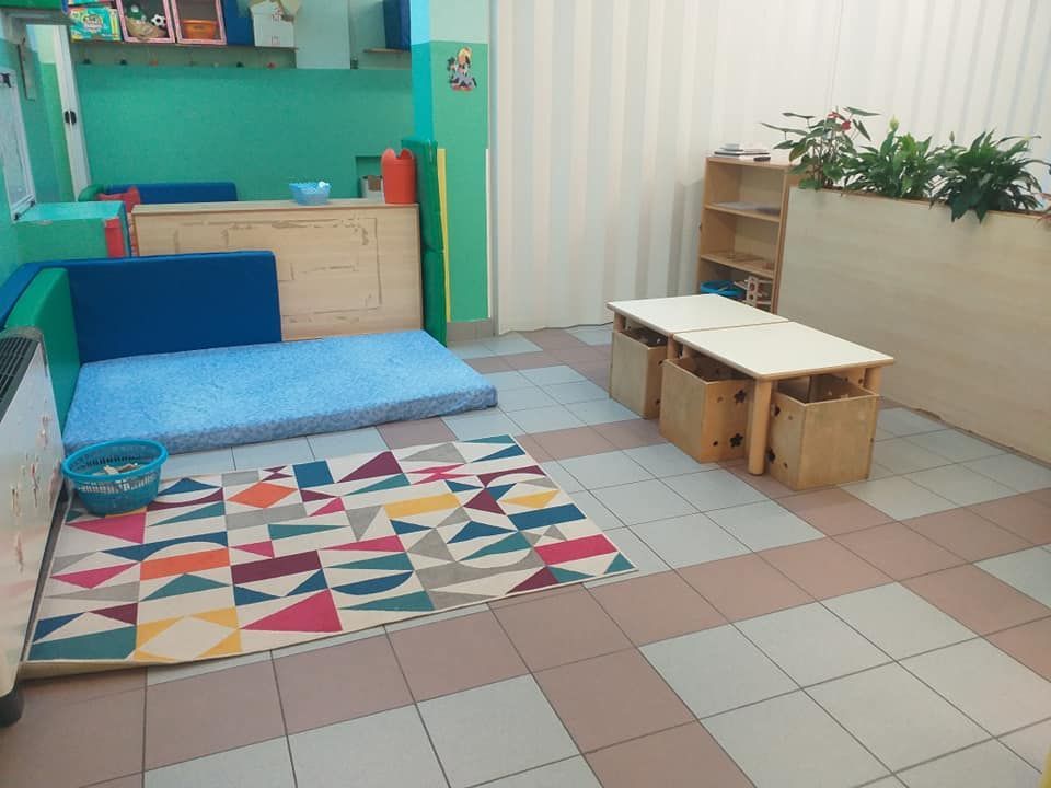 Stanza per l'infanzia: tappetino, tappeto, tavolo, scaffali, piante, pareti color verde acqua e pavimento in piastrelle color crema.