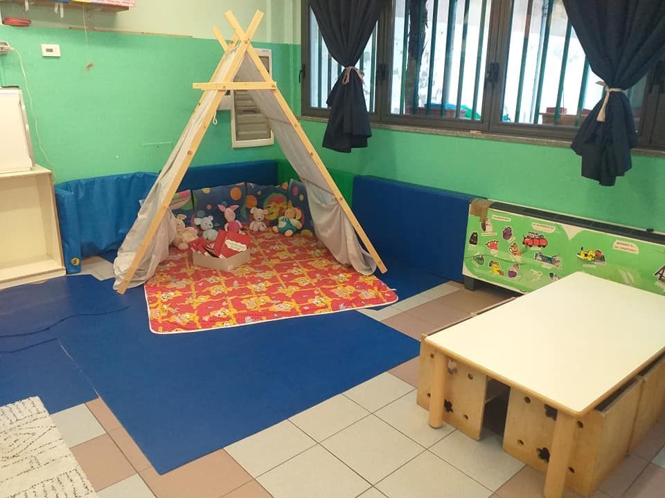 Stanza giochi per bambini con tenda indiana, tappetini, giocattoli e un tavolino. Le pareti sono verdi e blu.