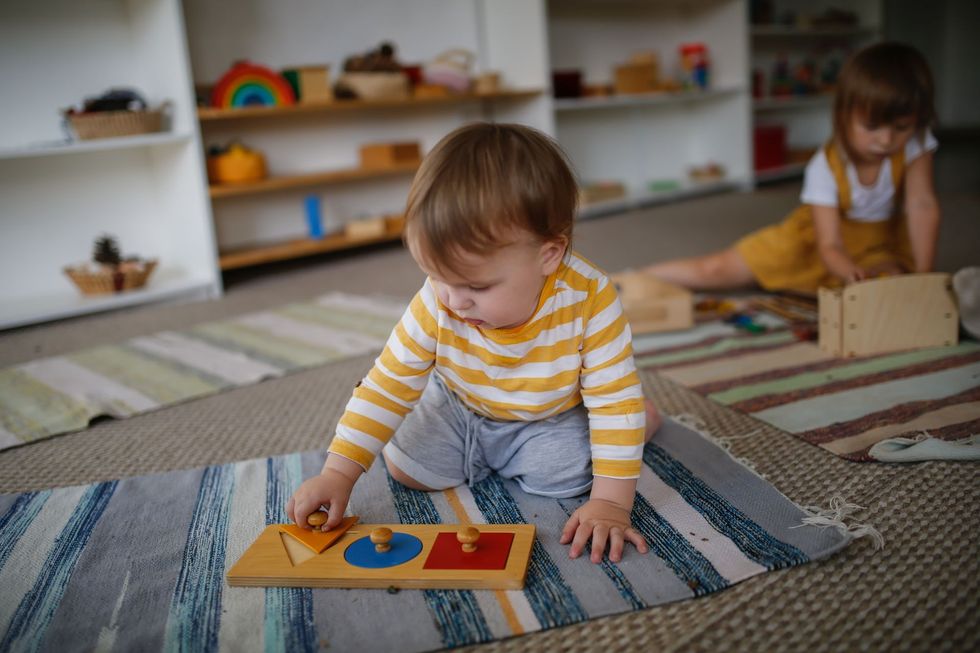 Bambino che gioca con un puzzle di forme su un tappeto in un'aula Montessori. Un altro bambino lavora sullo sfondo.