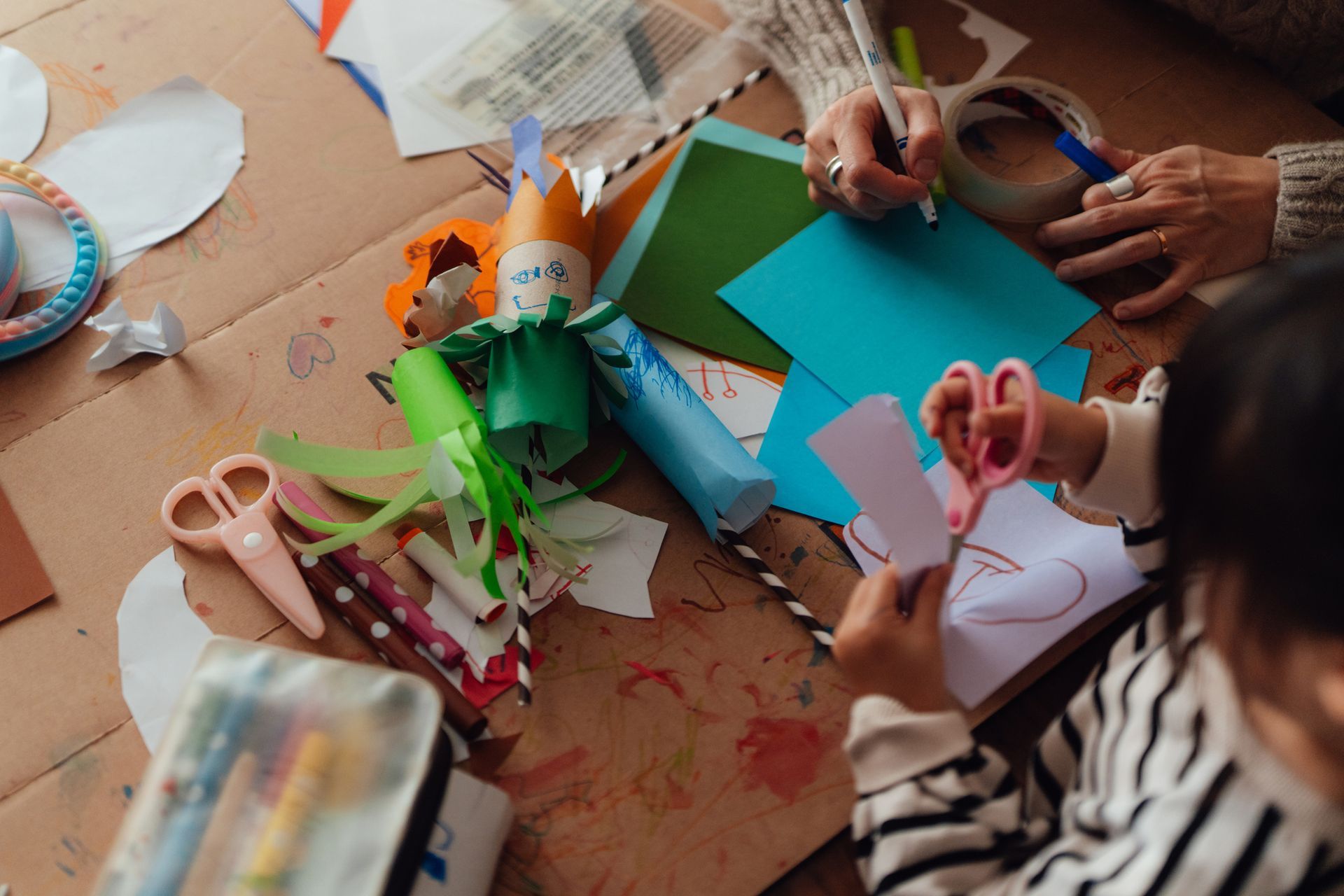 Bambini e adulti lavorano a un tavolo: disegnano con le penne e realizzano lavoretti colorati.
