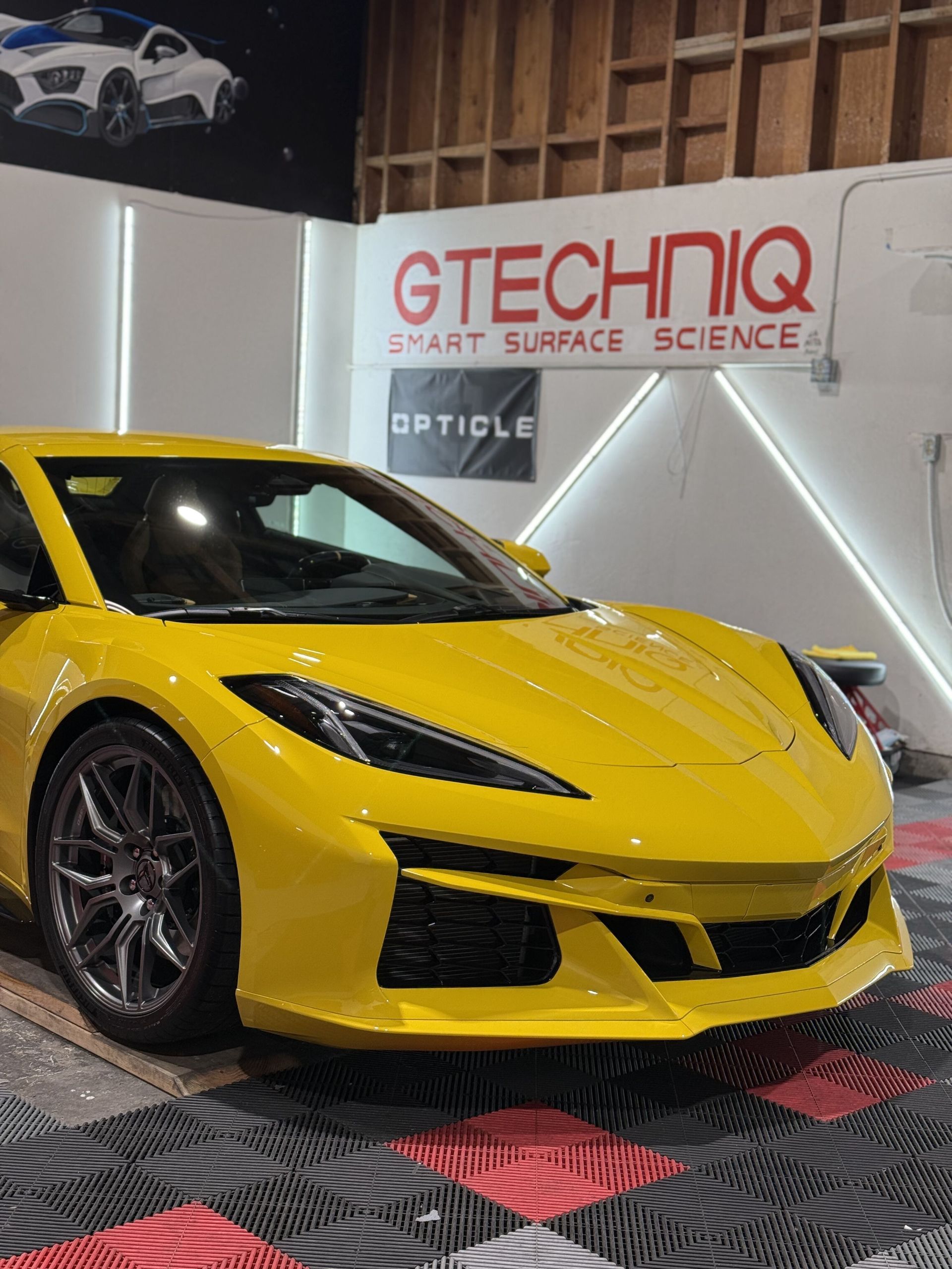A yellow sports car is parked in front of a sign that says opticle