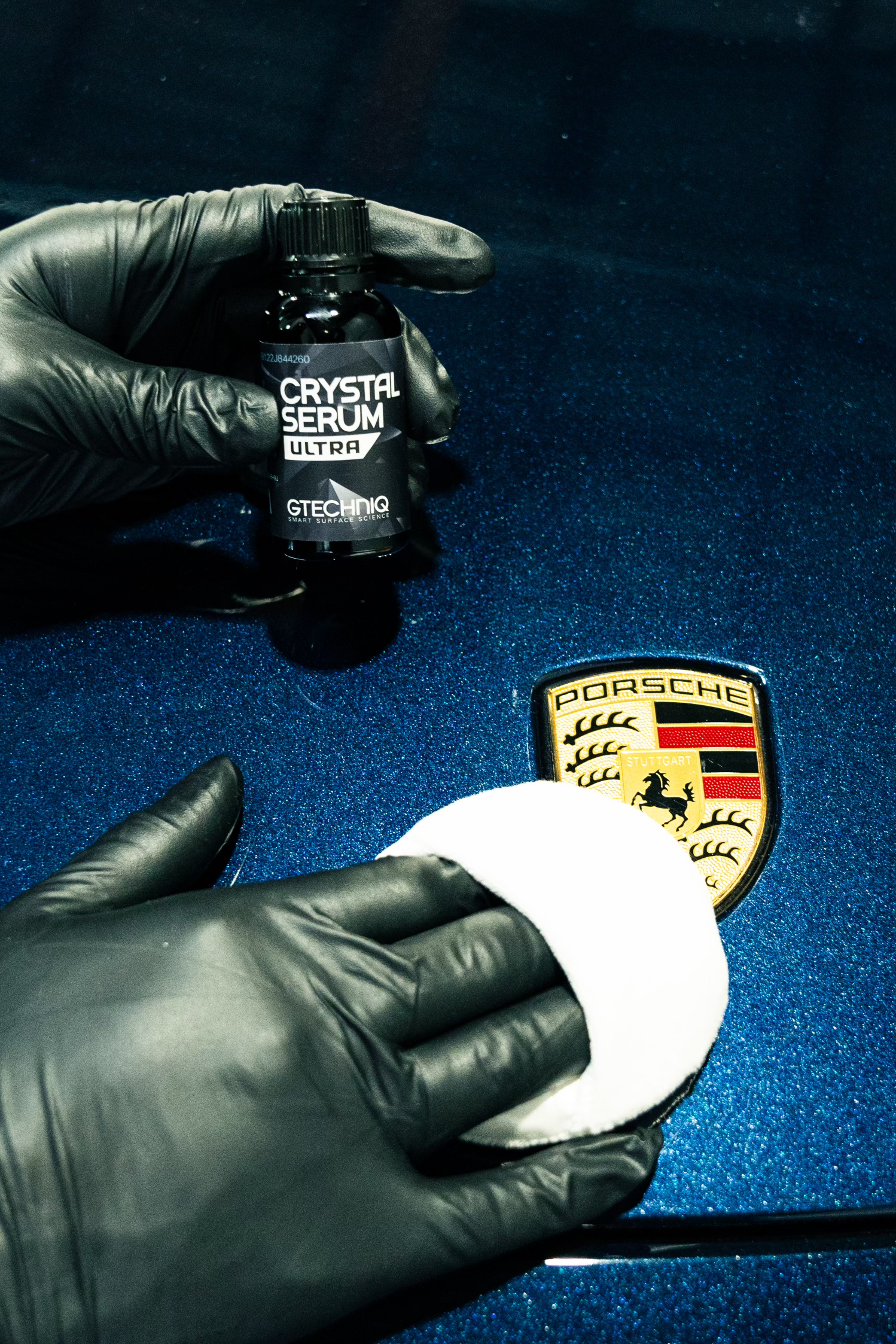 A person wearing black gloves is polishing a porsche logo on a blue car.