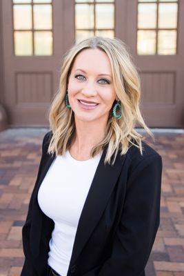 Smiling person in a black blazer and white top standing in front of a wooden building entrance