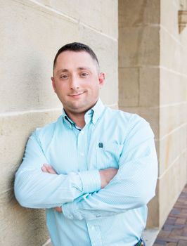 Man in a light blue striped shirt leaning against a stone wall with arms crossed