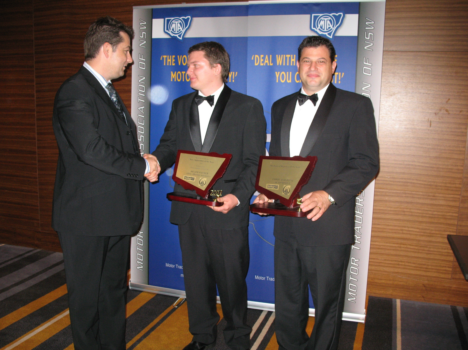 Man in suit shakes hands with two men in tuxedos holding plaques, against a blue banner backdrop.