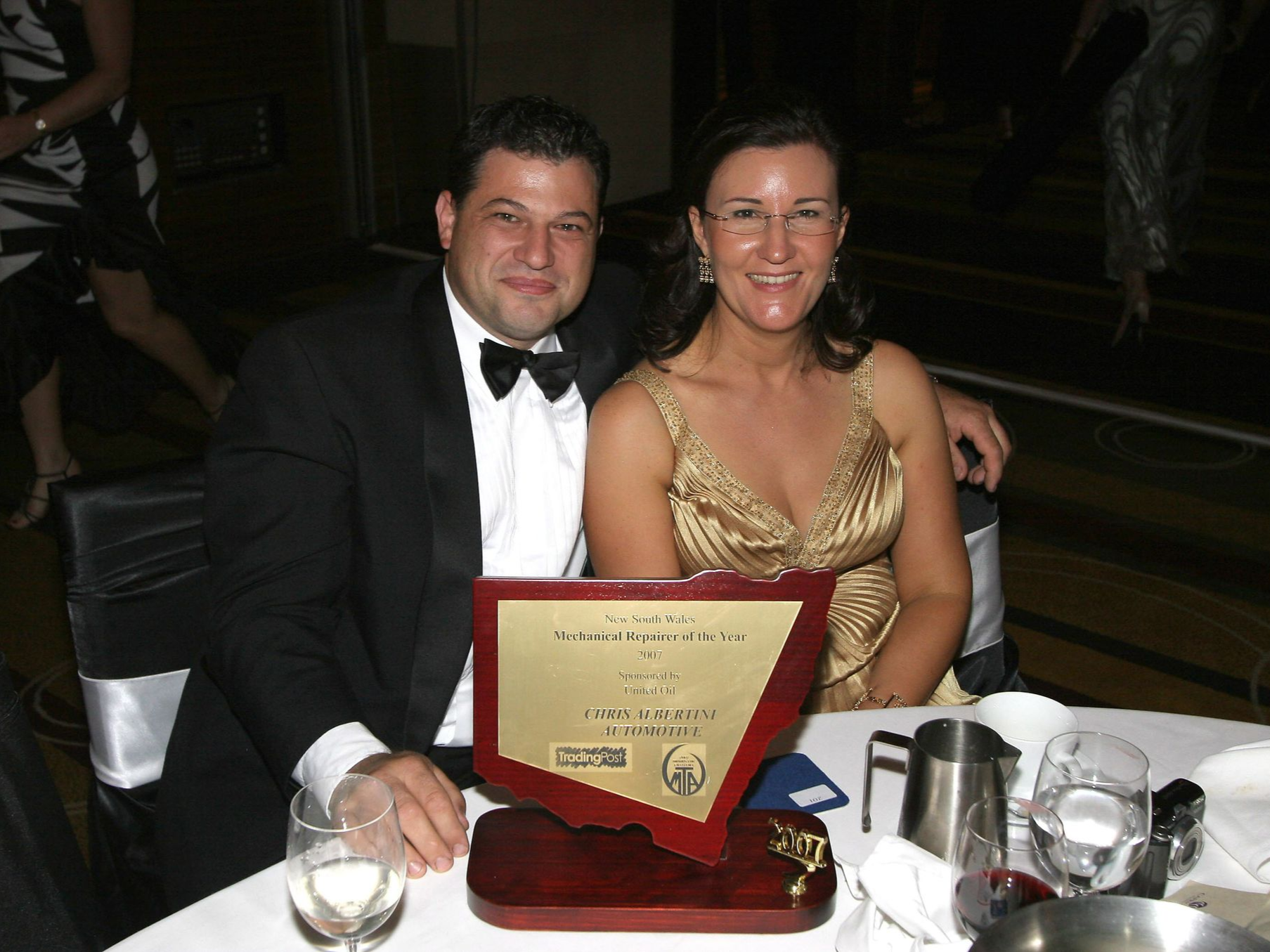 Couple at a formal event; holding a plaque. Man in a tuxedo, woman in a gold dress. Table with awards, silverware.