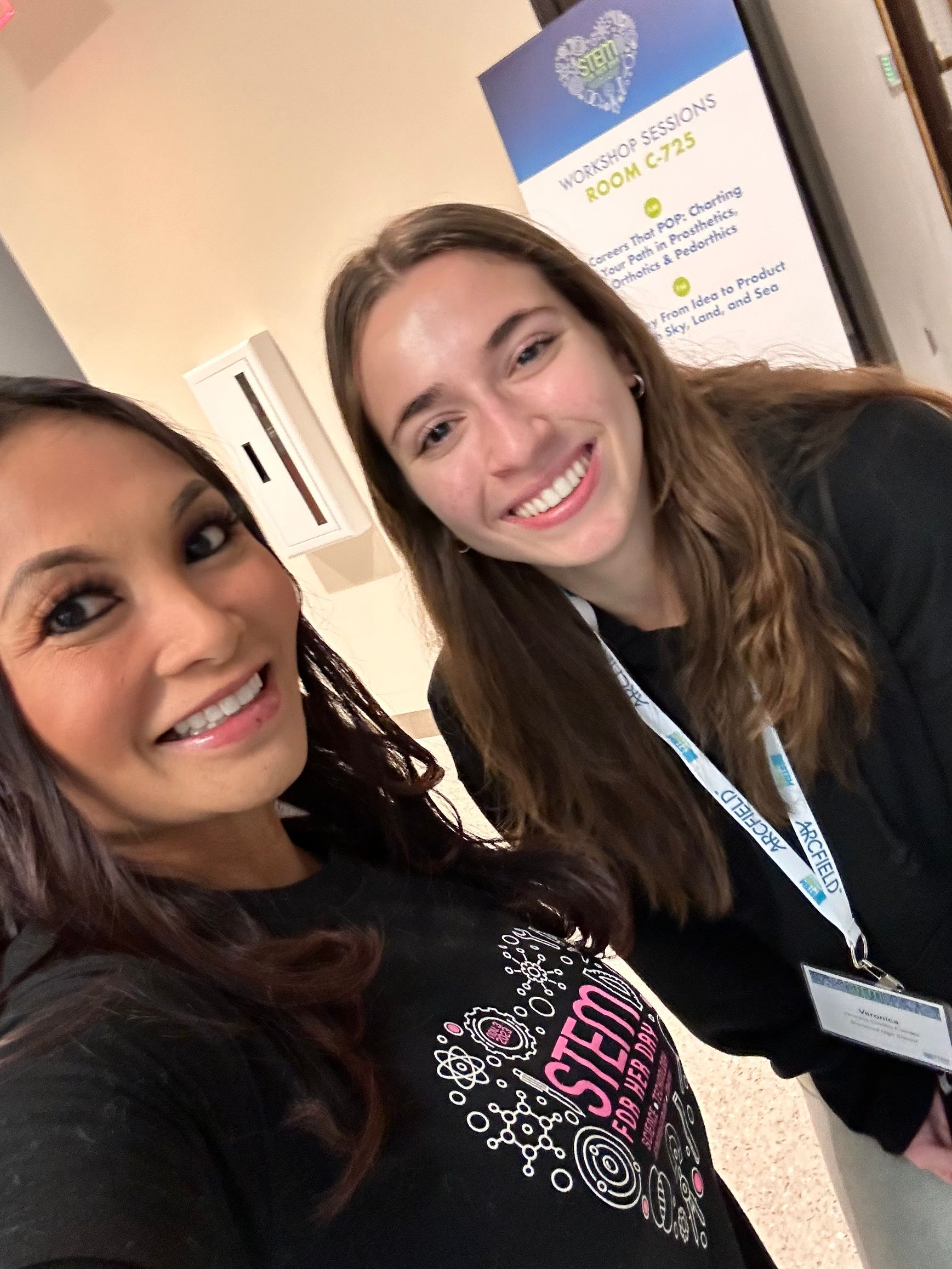 Two smiling women posing indoors at a conference, one wearing a lanyard.