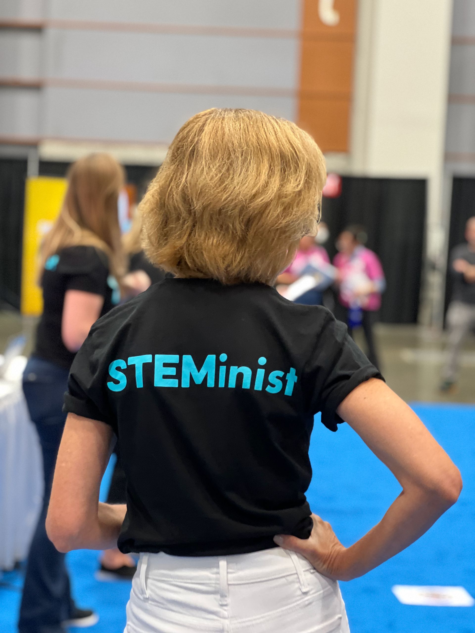 Person in a black STEMinist T-shirt stands with hands on hips at a conference or expo booth.