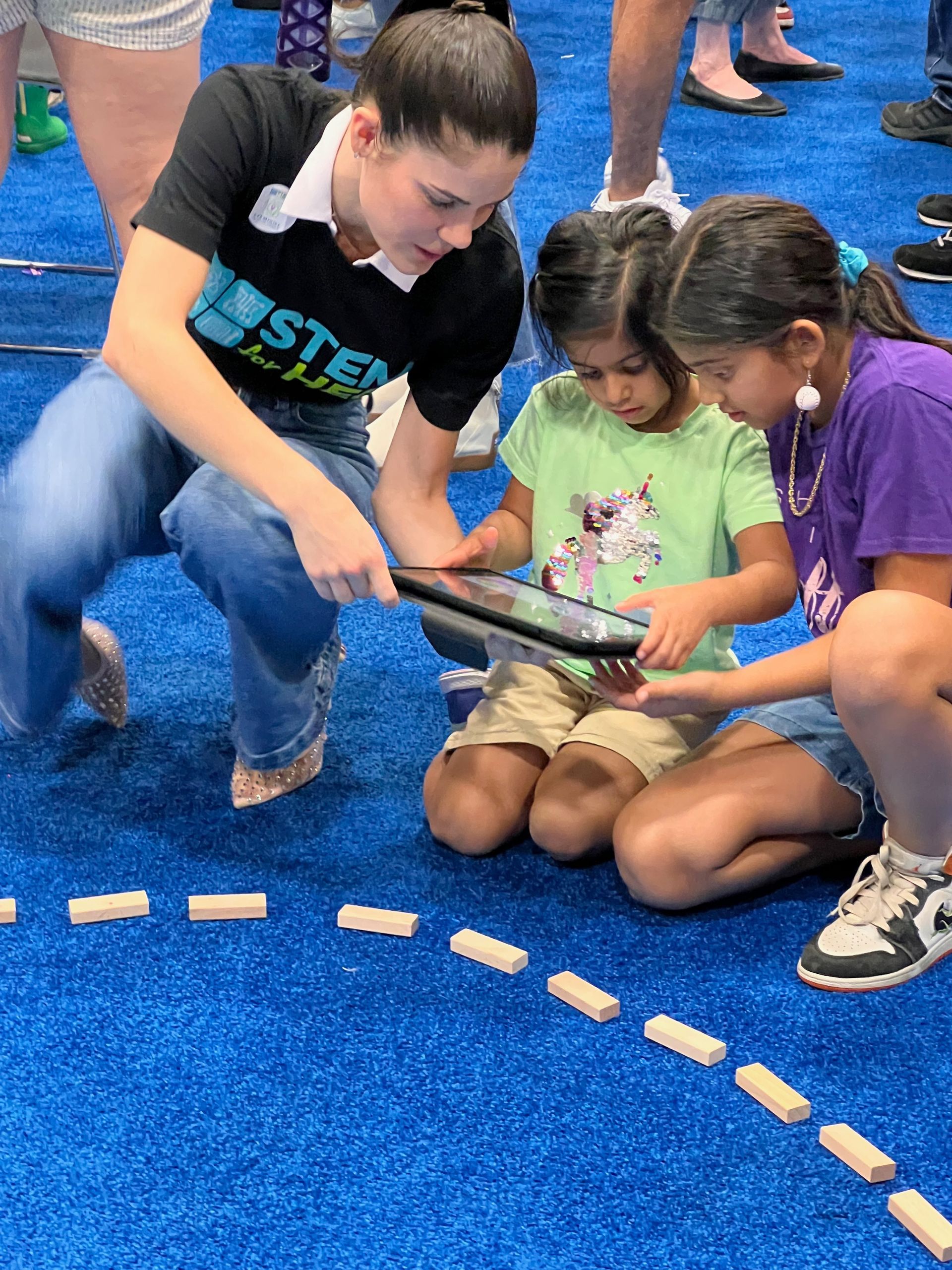 Children kneel on a blue floor, holding a tablet together in a collaborative activity.