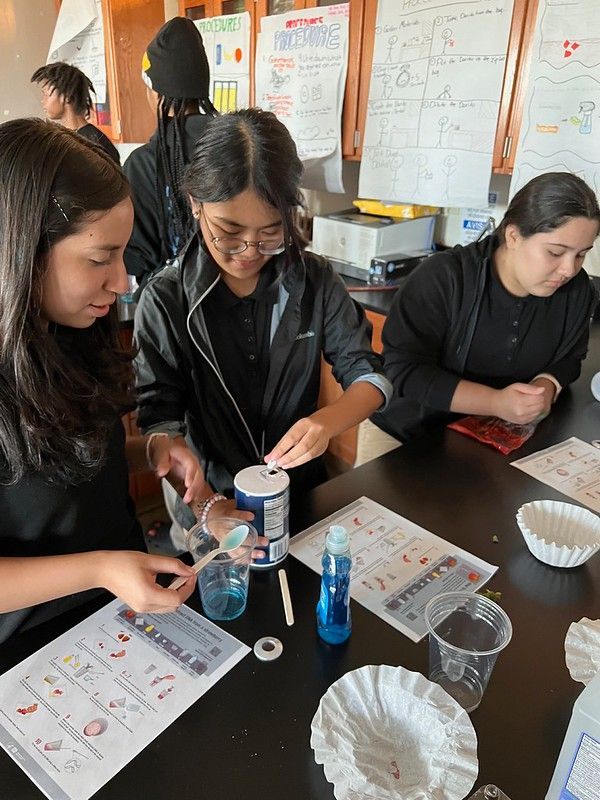 Three students at a table mixing materials in a classroom science activity.