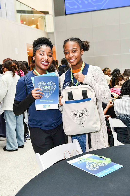 Two smiling people hold blue brochures at an indoor event table.