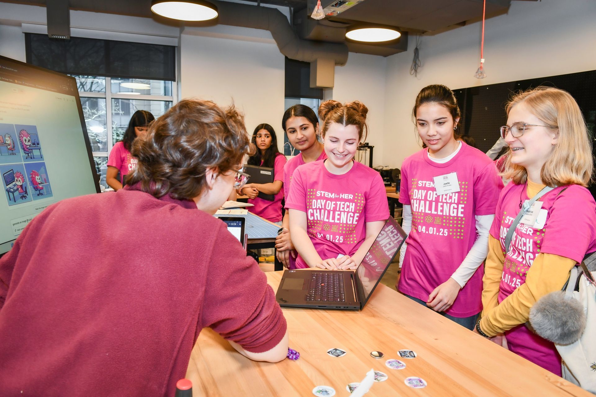 Group in pink shirts gathered around a table, listening to a man in a maroon shirt indoors.