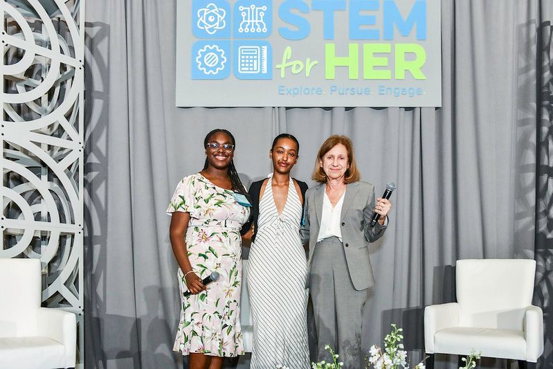 Three women pose onstage beneath a STEM for HER banner, smiling and holding bouquets.