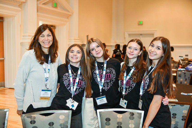 Five women standing together indoors at a conference, smiling with name badges and backpacks
