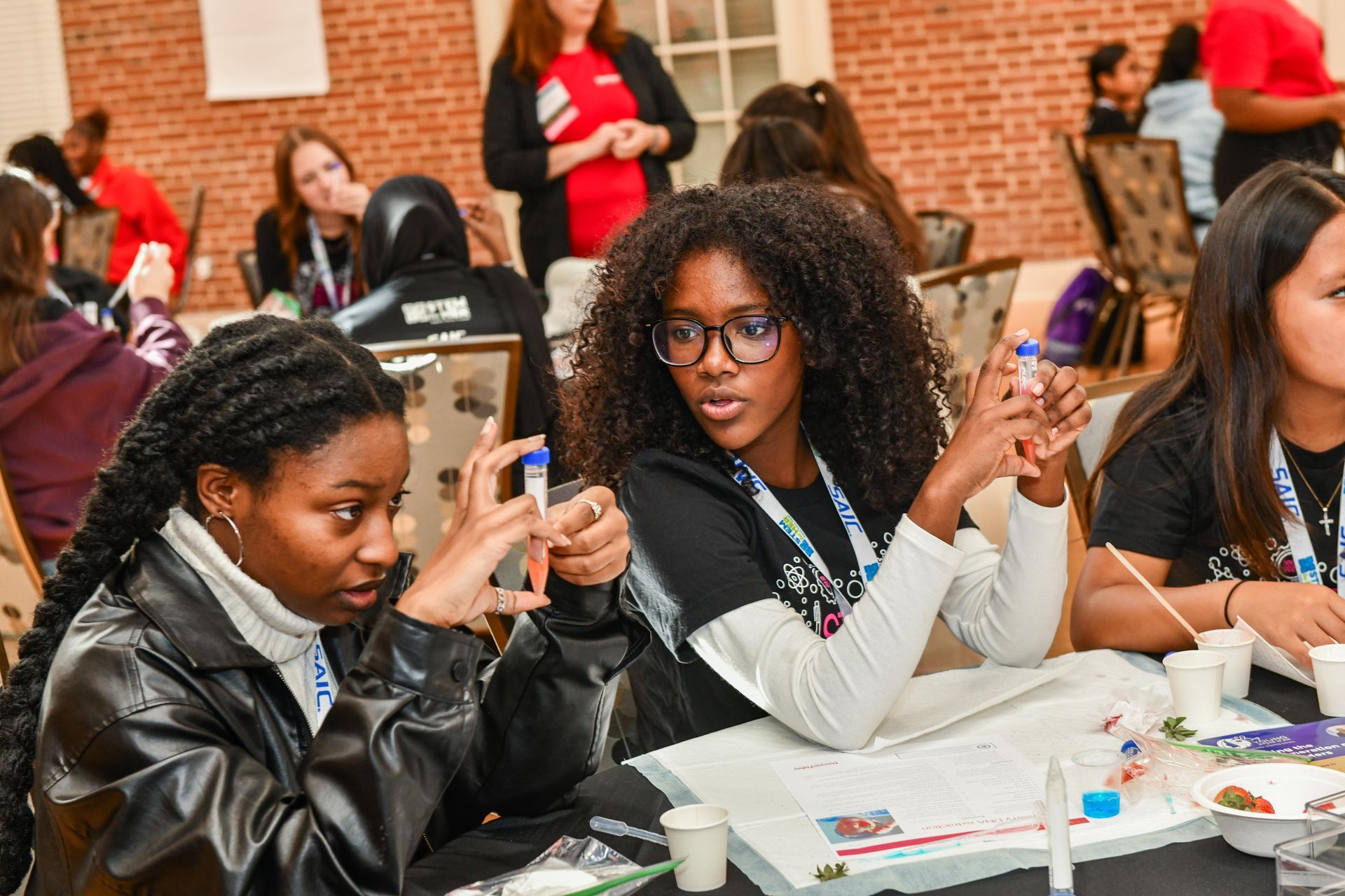 Students seated at a table, using smartphones and talking in a classroom or event space
