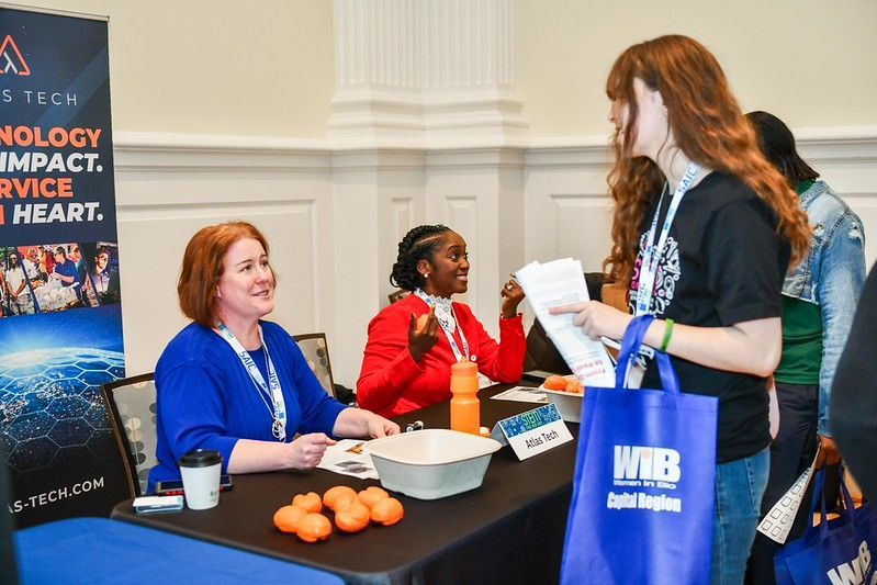 Two women at a table handing out information to a visitor at a conference booth with brochures and oranges.