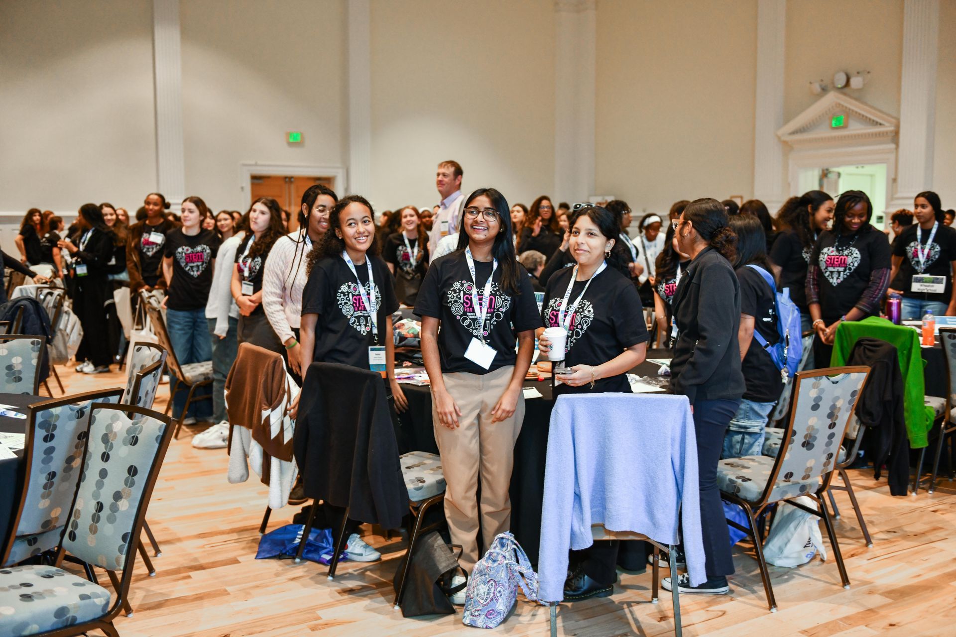 Conference attendees standing and chatting in a ballroom beside round tables and chairs