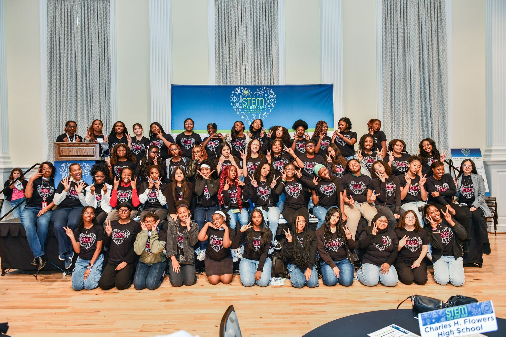 Large group posing on a stage in front of a blue backdrop and curtains, many wearing black shirts and jeans.