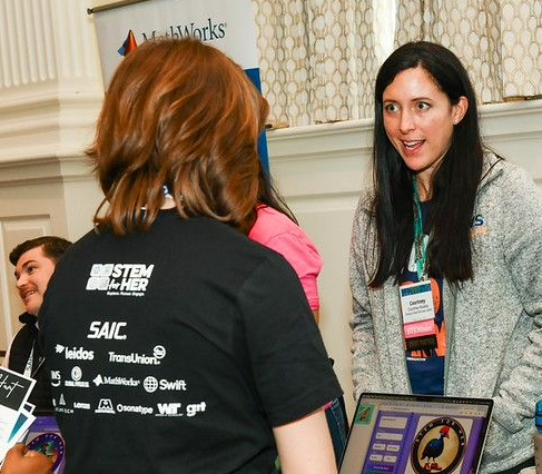 Two people converse at a conference table with posters, badges, and a laptop in a bright room.