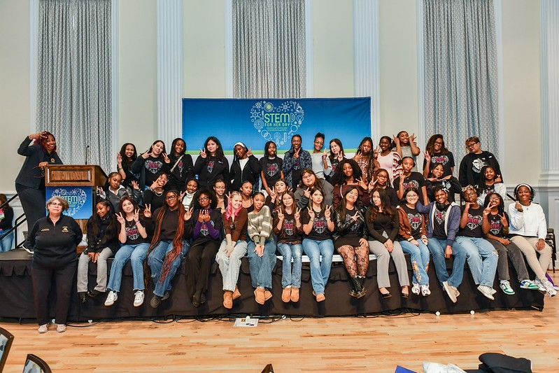 Large group posing on a stage in front of blue curtains and a banner, seated and standing together.