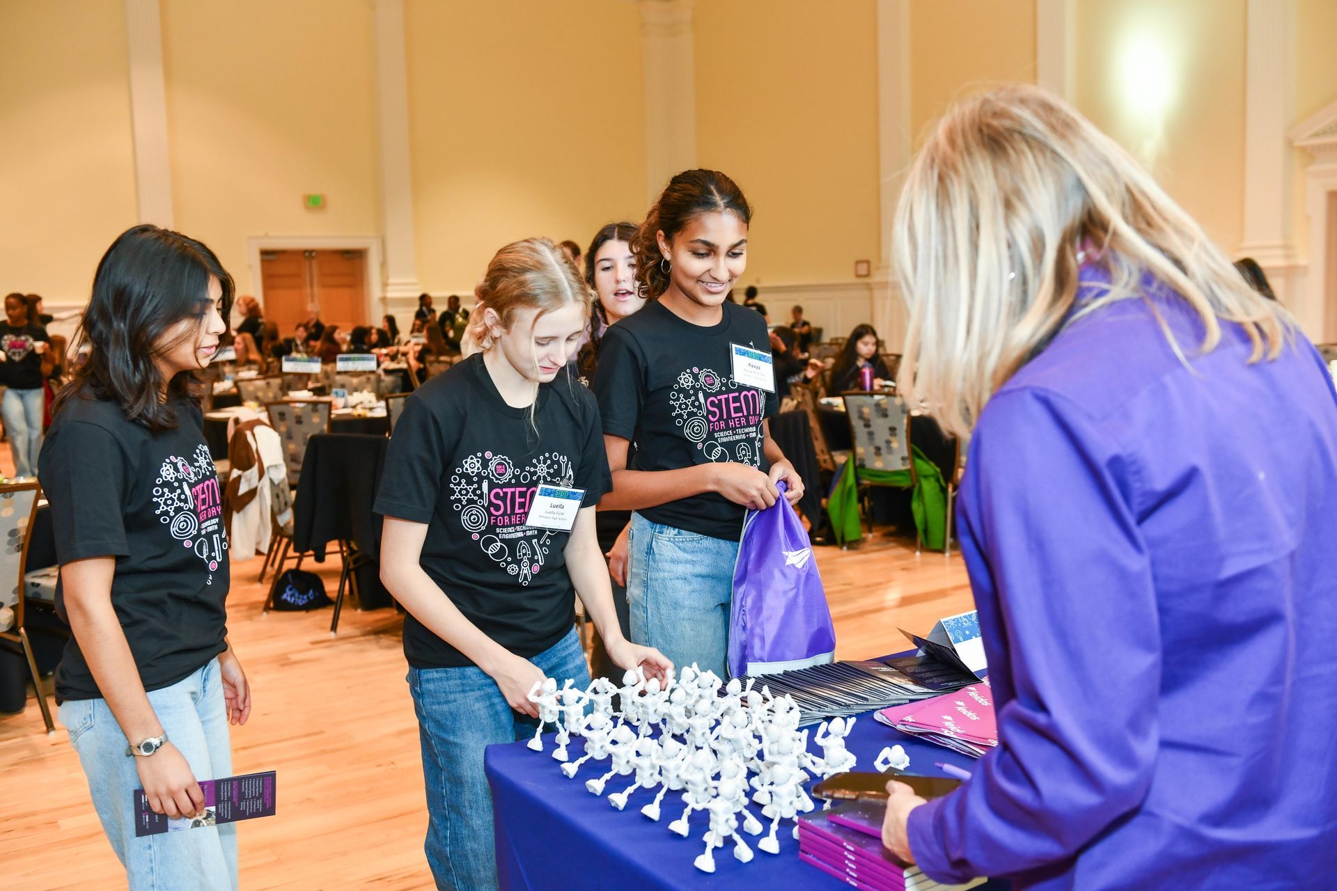 Students leaning over a table of chess pieces while a woman in a purple shirt watches at a competition table.