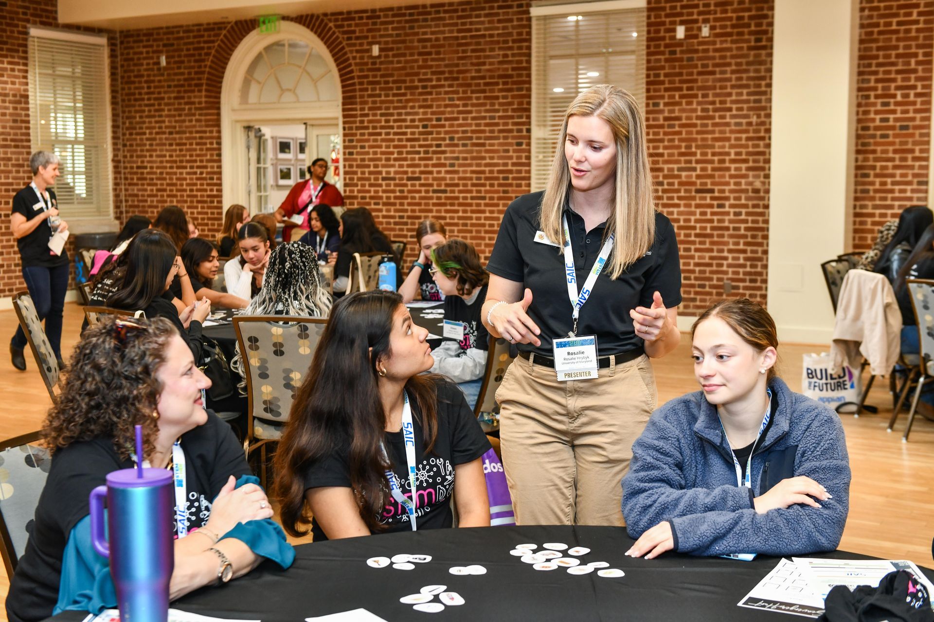 People gathered around a table at an indoor event, chatting over cards and water bottles