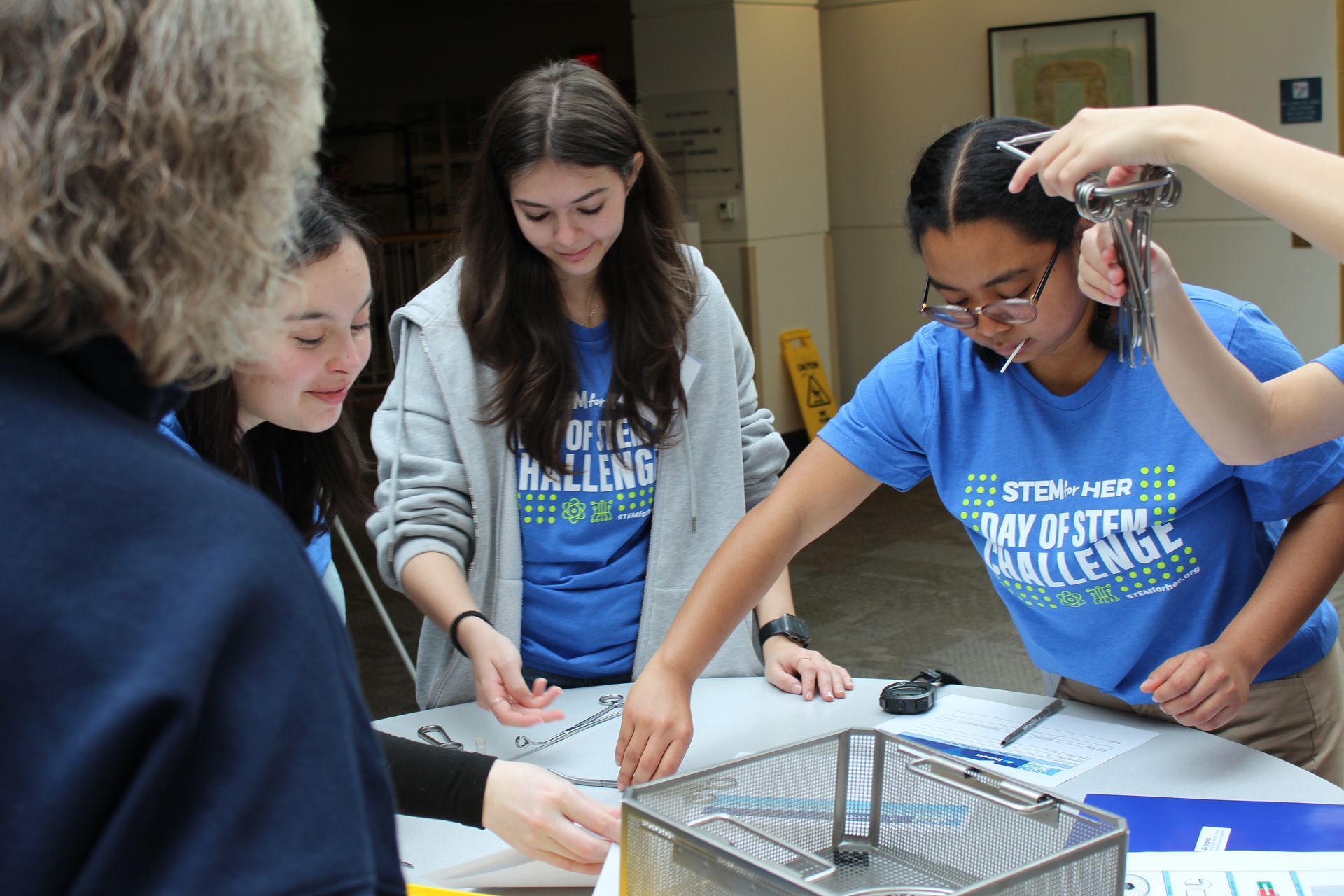 Three people in blue shirts collaborate around a table with papers and tools in a classroom.